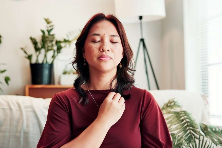 Une femme assise les yeux fermés, une main sur la poitrine, pratiquant des techniques de respiration consciente dans son salon.